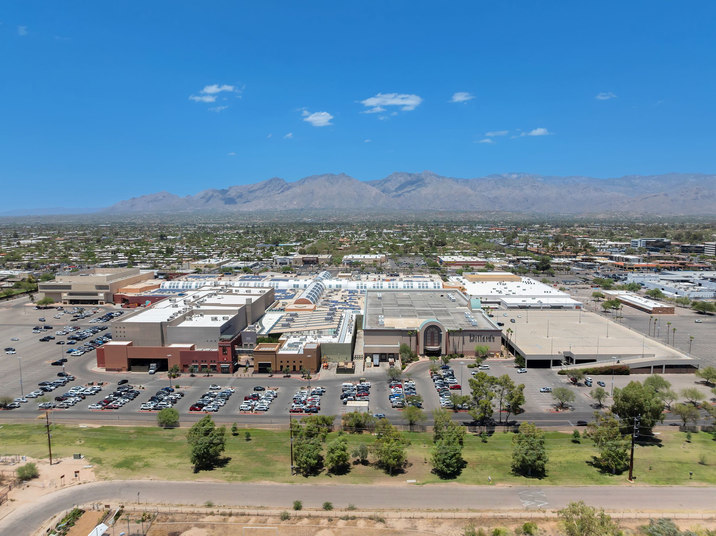 Aerial view of a large shopping mall complex with parking lots, surrounded by urban sprawl and mountain backdrop.