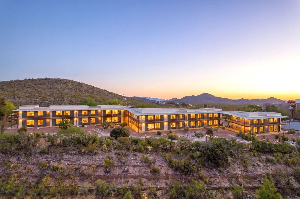 A modern desert hotel glows warmly at dusk, nestled among rugged hills under a stunning gradient sunset sky.