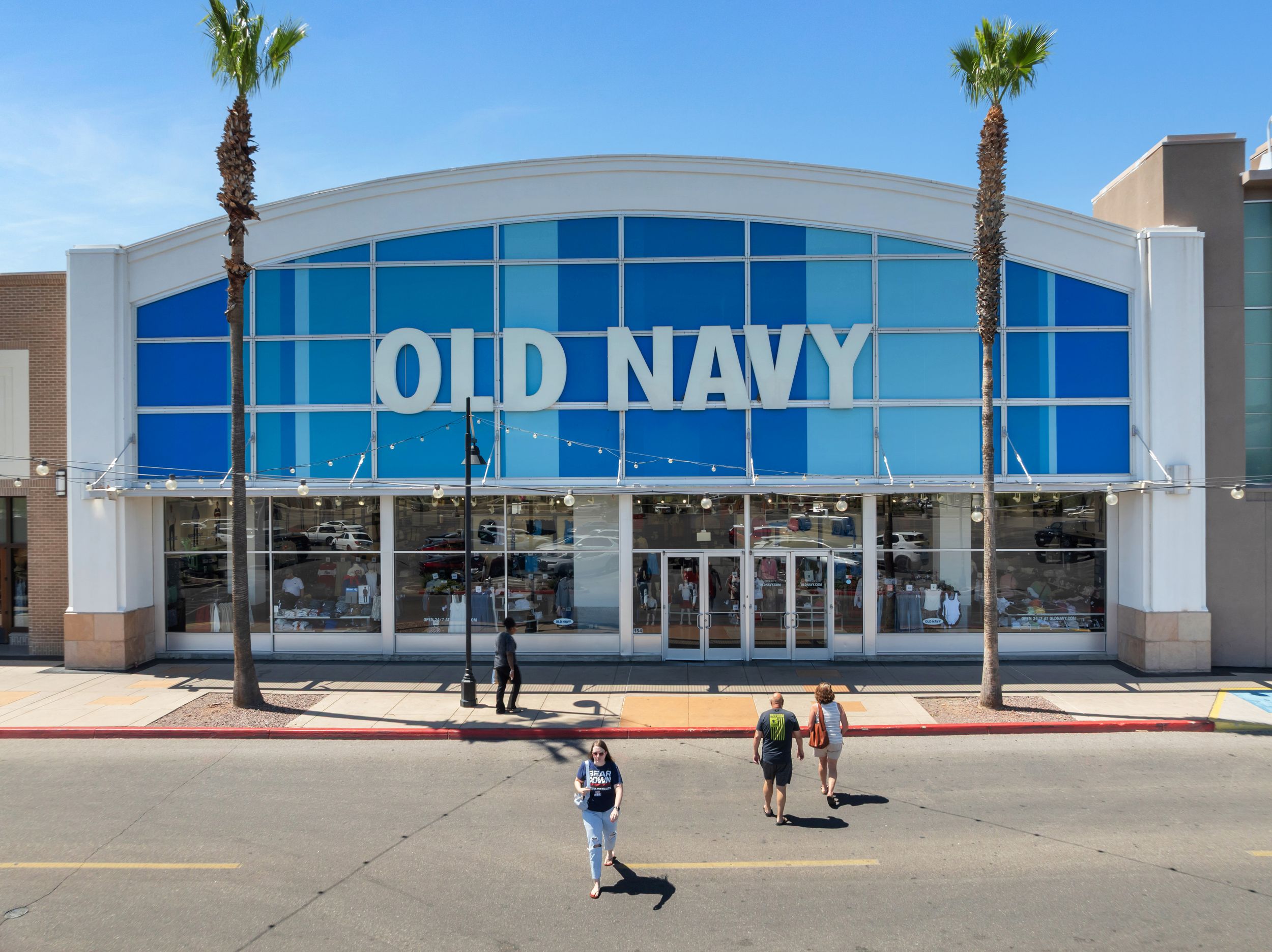 Old Navy retail store exterior with large blue facade, palm trees, and shoppers walking in a sunny parking lot.