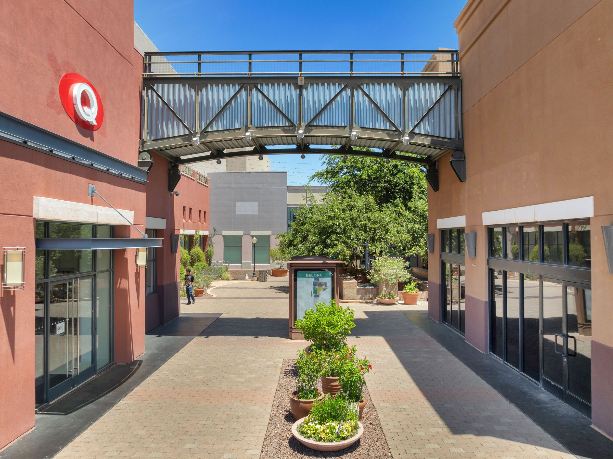 Outdoor shopping plaza walkway with Target store, pedestrian bridge, potted plants, and clear blue sky.