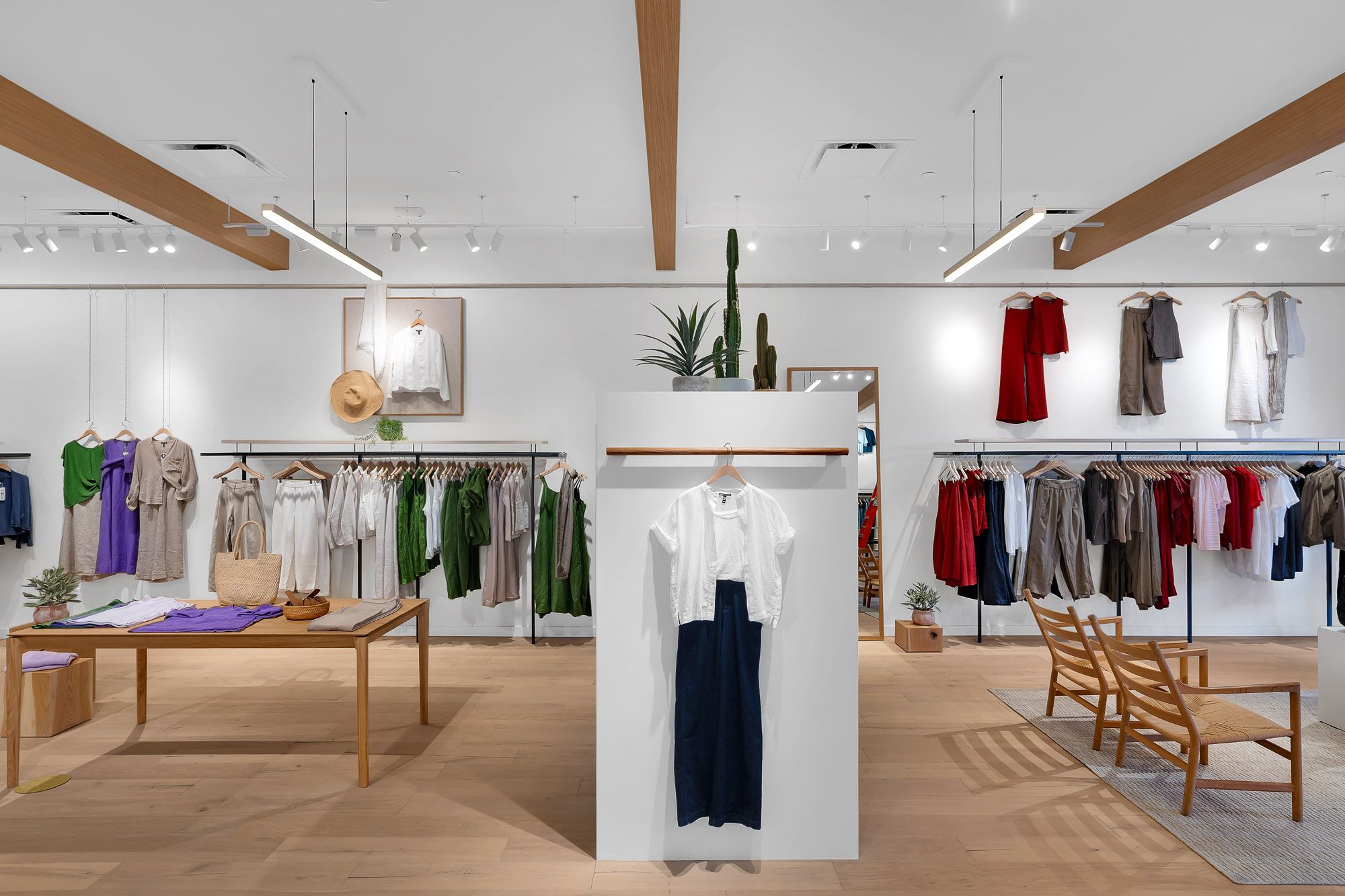 Bright clothing store interior featuring wooden display tables, hanging garments in various colors, and cactus plant decor.