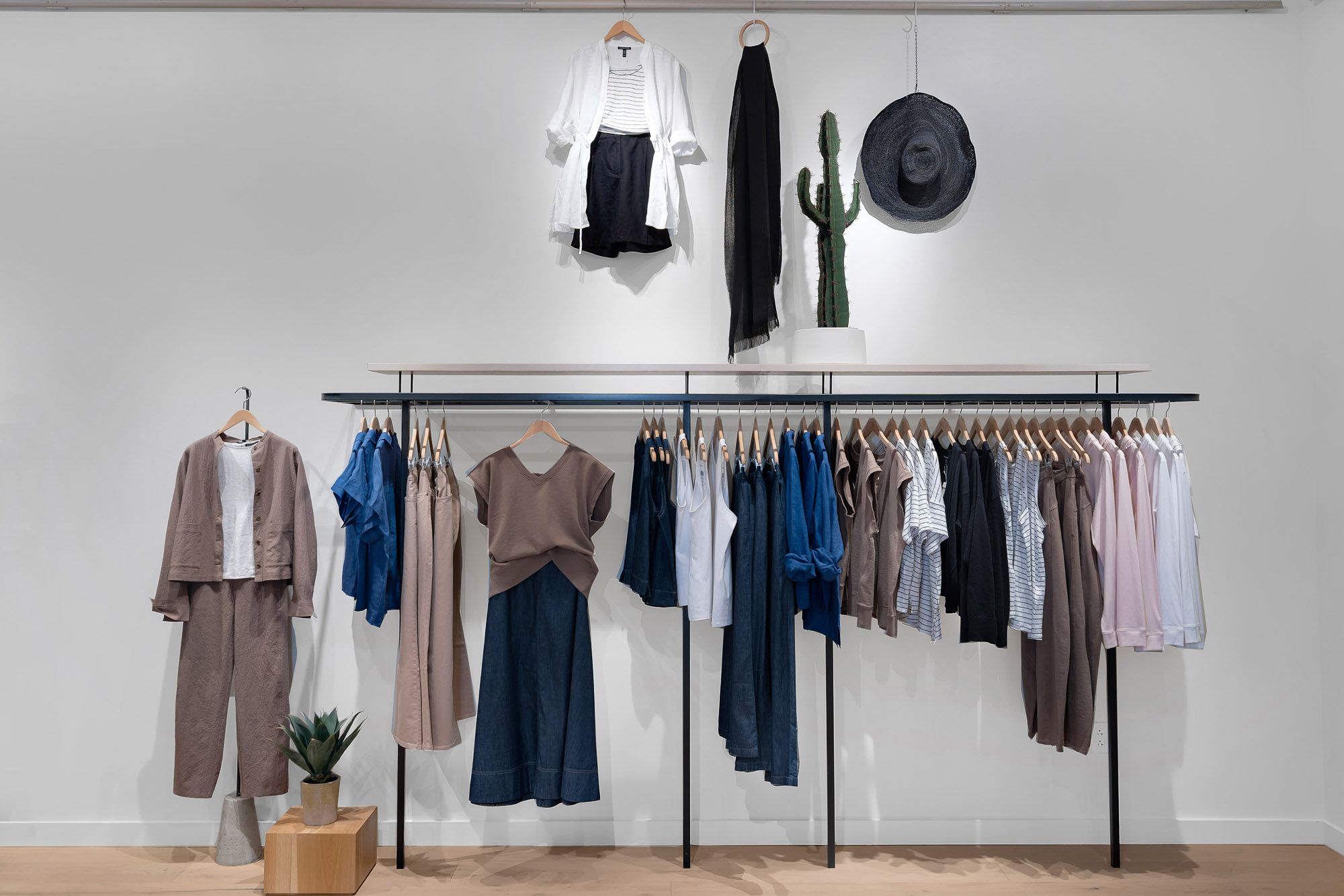 Modern clothing boutique displaying neutral-toned apparel on black metal racks with a cactus decor and hat wall display.