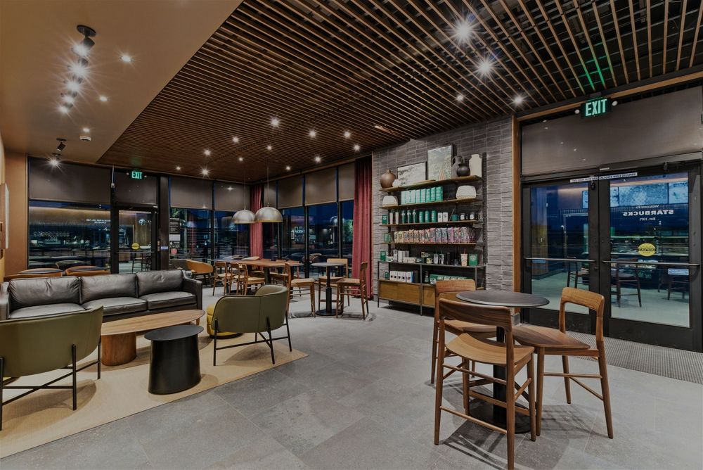 Contemporary cafe seating area with wooden tables, comfortable chairs, and retail shelving against dark walls.