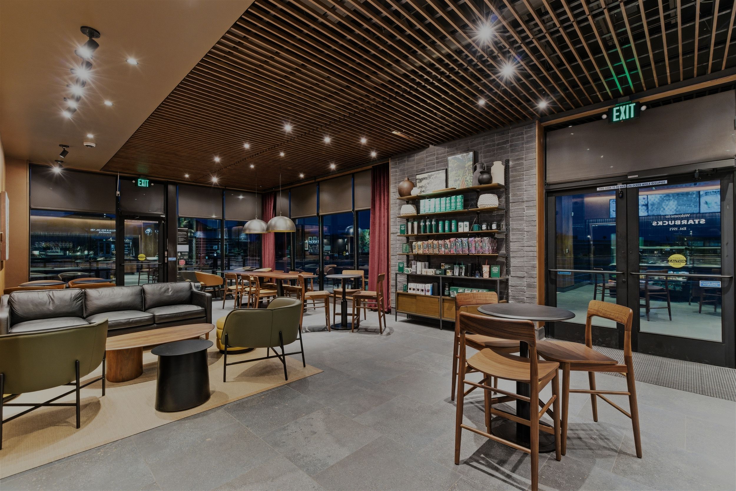 Contemporary cafe seating area with wooden tables, comfortable chairs, and retail shelving against dark walls.