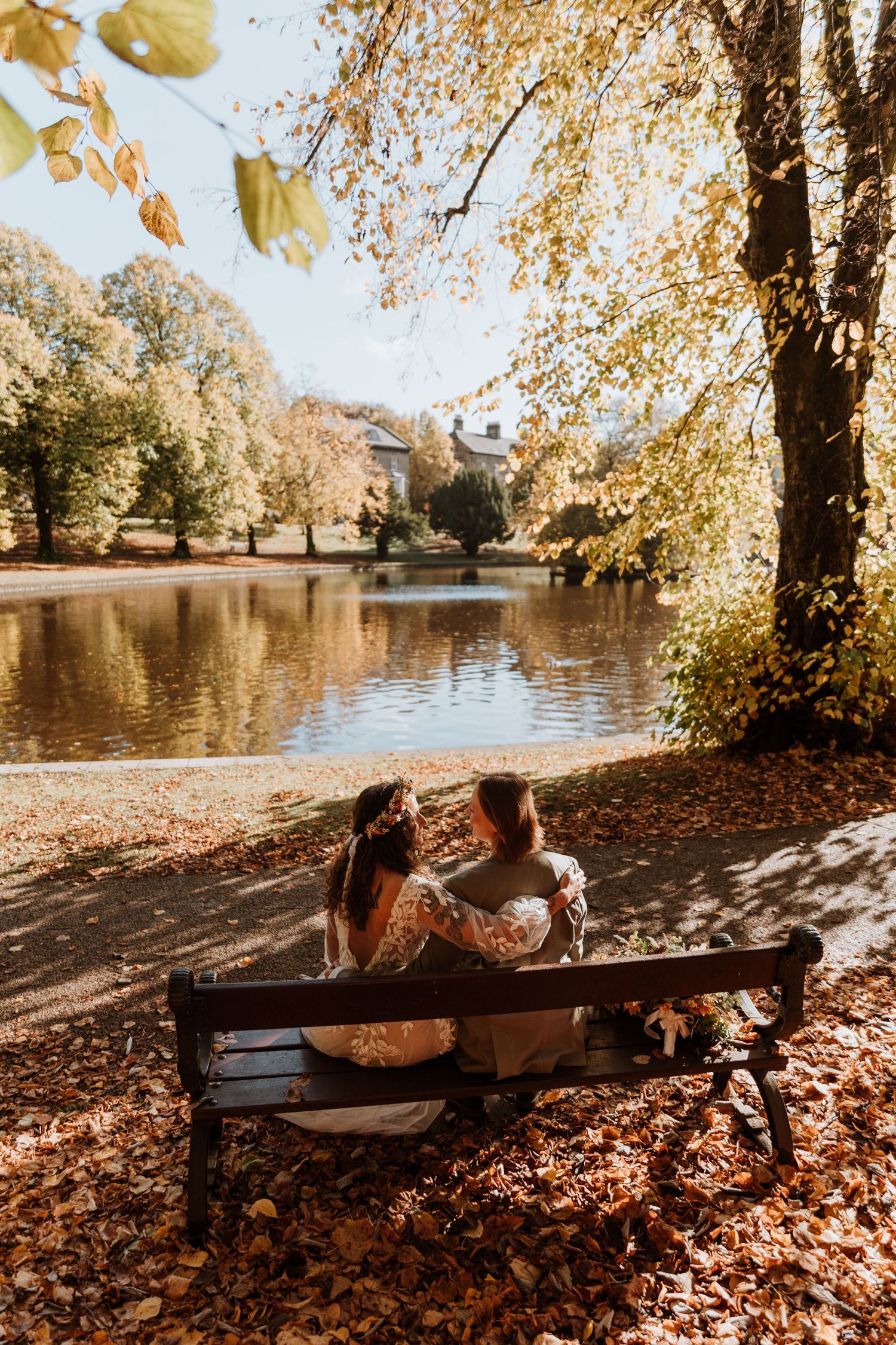 A couple relaxes on a wooden bench by a lake surrounded by autumn trees with golden leaves falling on the ground at Buxton Park.