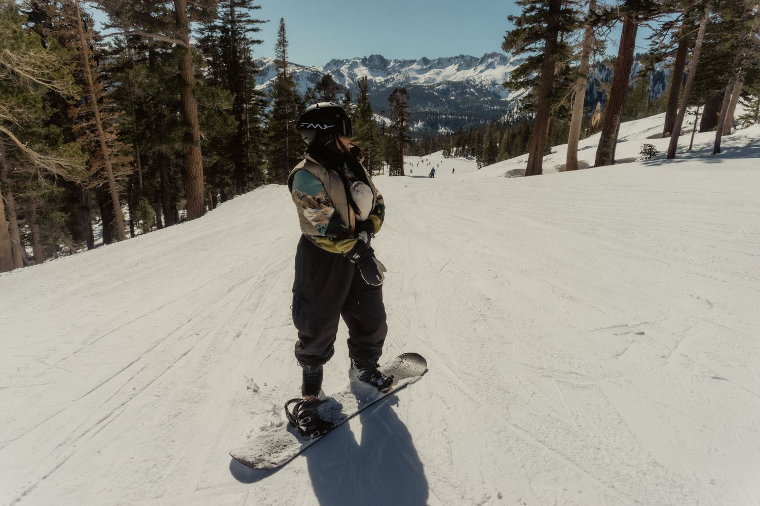 A snowboarder performs a jump sequence on a snowy slope with evergreen trees and mountains in the background.