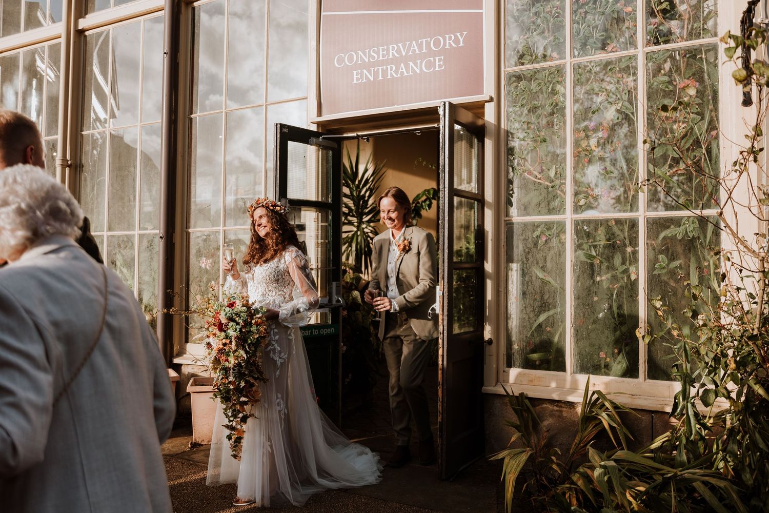 Wedding party exits through glass doors of a conservatory with elegant floral decorations and natural lighting.
