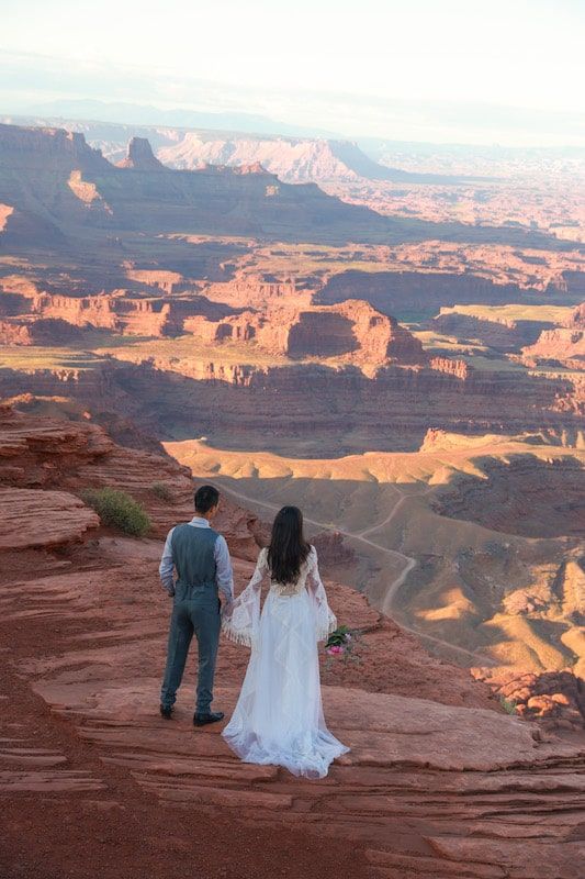 A couple sits on red rocks overlooking the vast canyons and mesas of Dead Horse Point State Park at sunset.