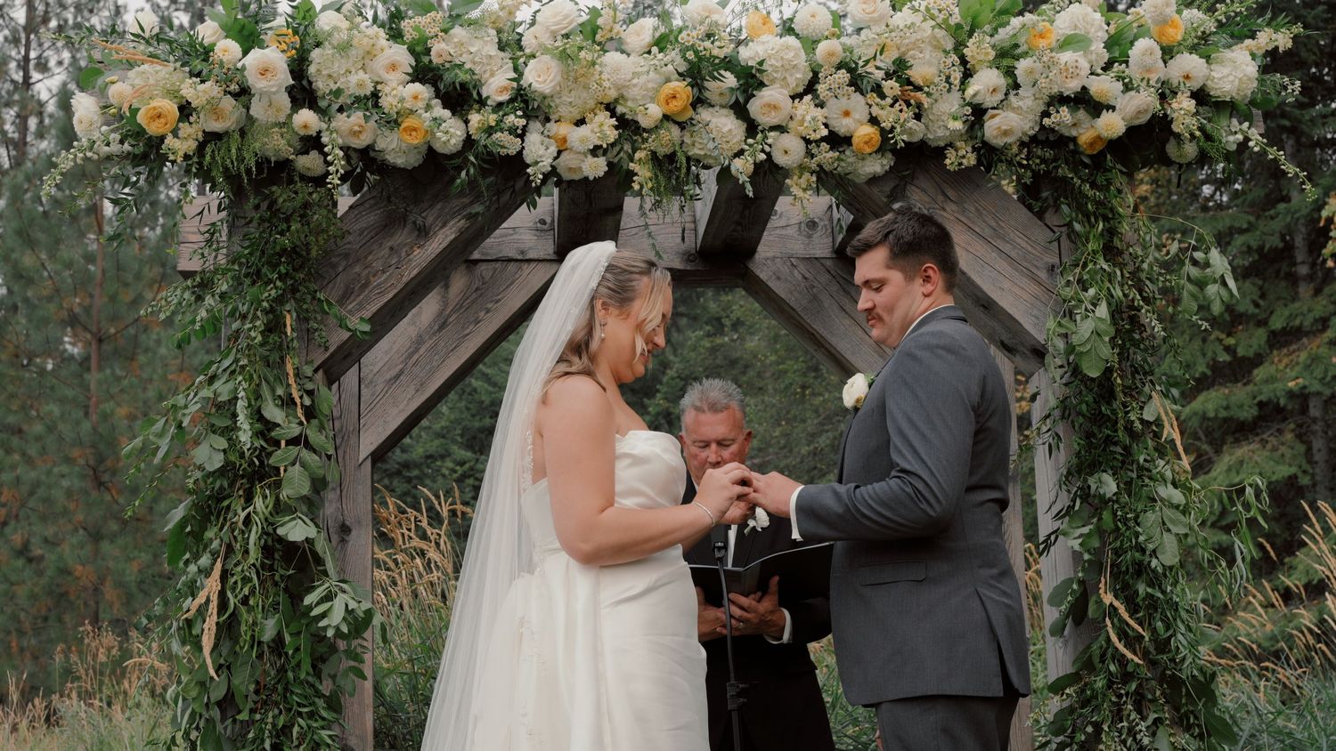 A couple exchanges wedding vows under a floral arch decorated with white roses and greenery during an outdoor ceremony.