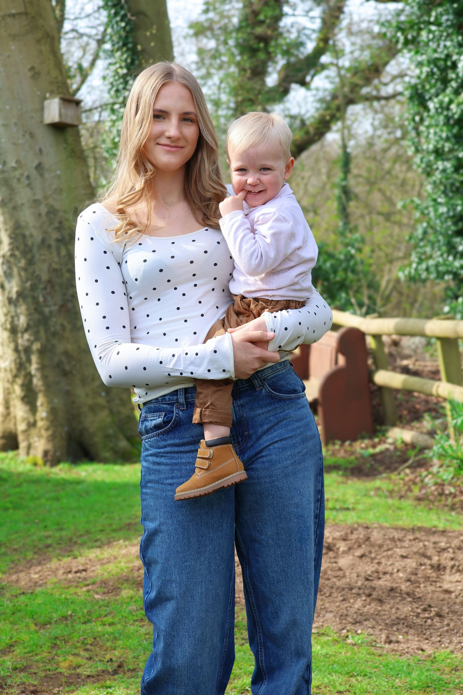 A baby wearing a cream knit vest and olive shorts sits on a textured blanket outdoors in a grassy field.