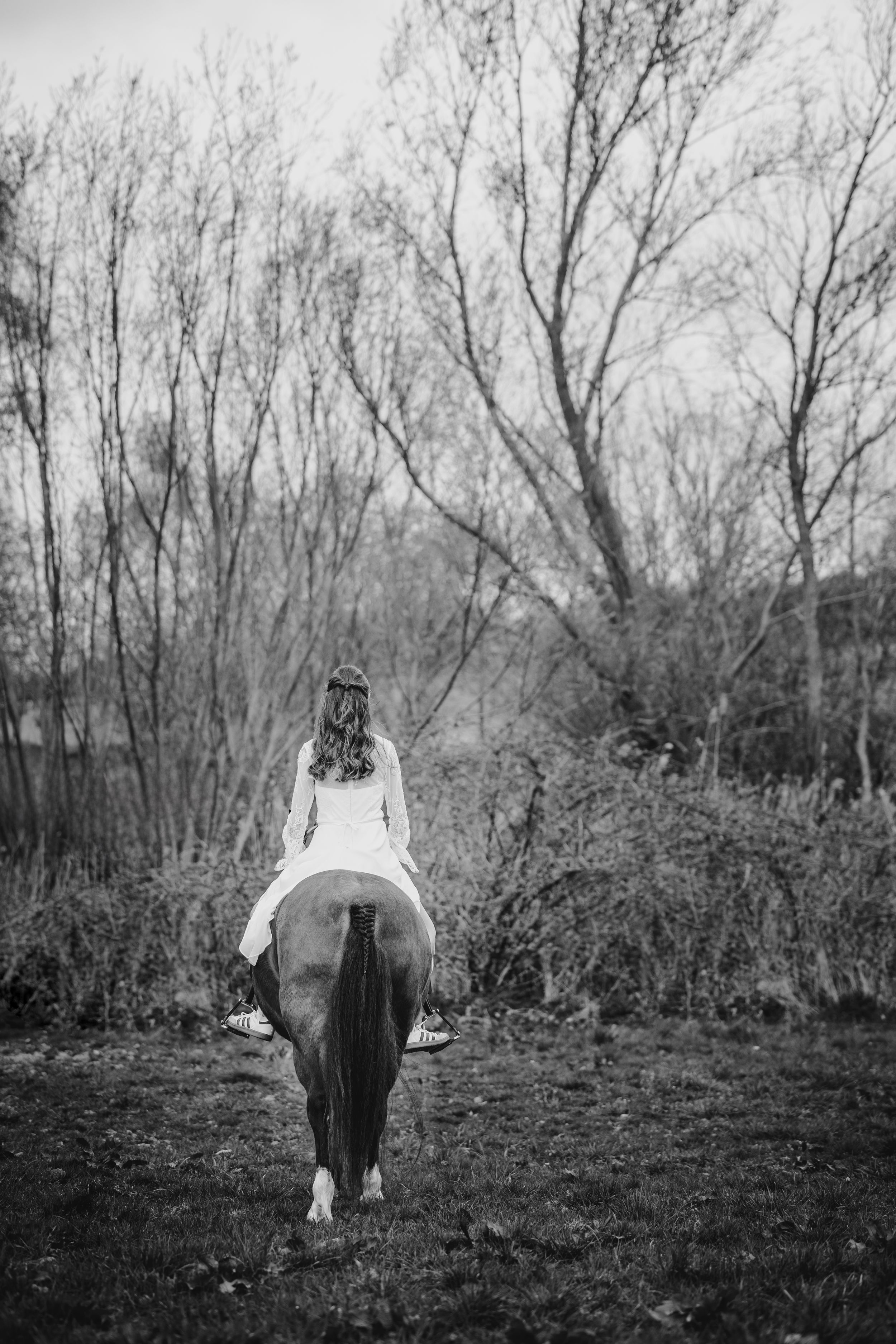 Black and white photo of a woman in a white dress riding a horse bareback through a wooded landscape.