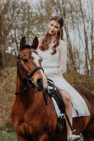A smiling young woman in a white lace dress sits atop a chestnut horse in a forest setting.