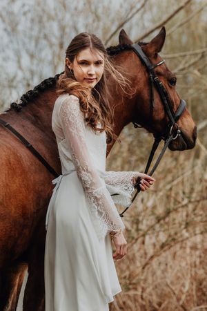 Blonde woman in elegant white lace bridal gown standing beside a bay horse in a natural outdoor setting.