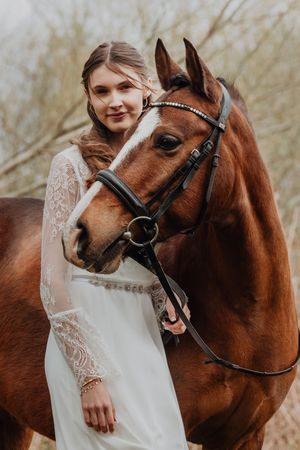 Bride in white lace dress posing with a chestnut horse wearing a bridle outdoors.