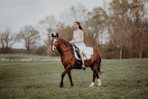 Young woman in white dress riding a chestnut horse across a green field on a cloudy day.
