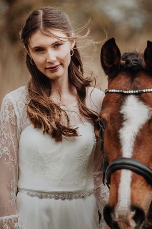 A smiling bride in a white lace wedding gown poses beside a chestnut horse with a bridle outdoors.