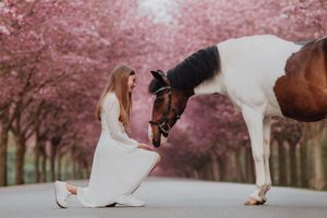 Woman in white dress kneeling beside a brown and white horse among pink cherry blossom trees.