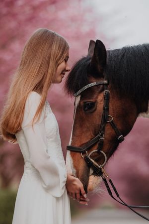 A blonde woman in a white dress stands face-to-face with a bay horse amid pink cherry blossom trees.
