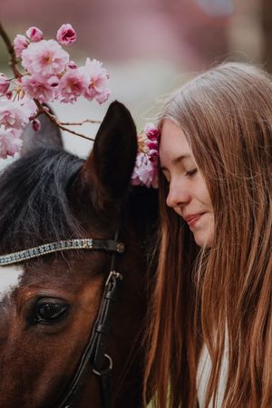 Young woman with long hair resting forehead against brown horse adorned with pink cherry blossoms.