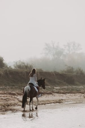 Woman in white dress riding a horse along a misty shoreline with trees in the background.