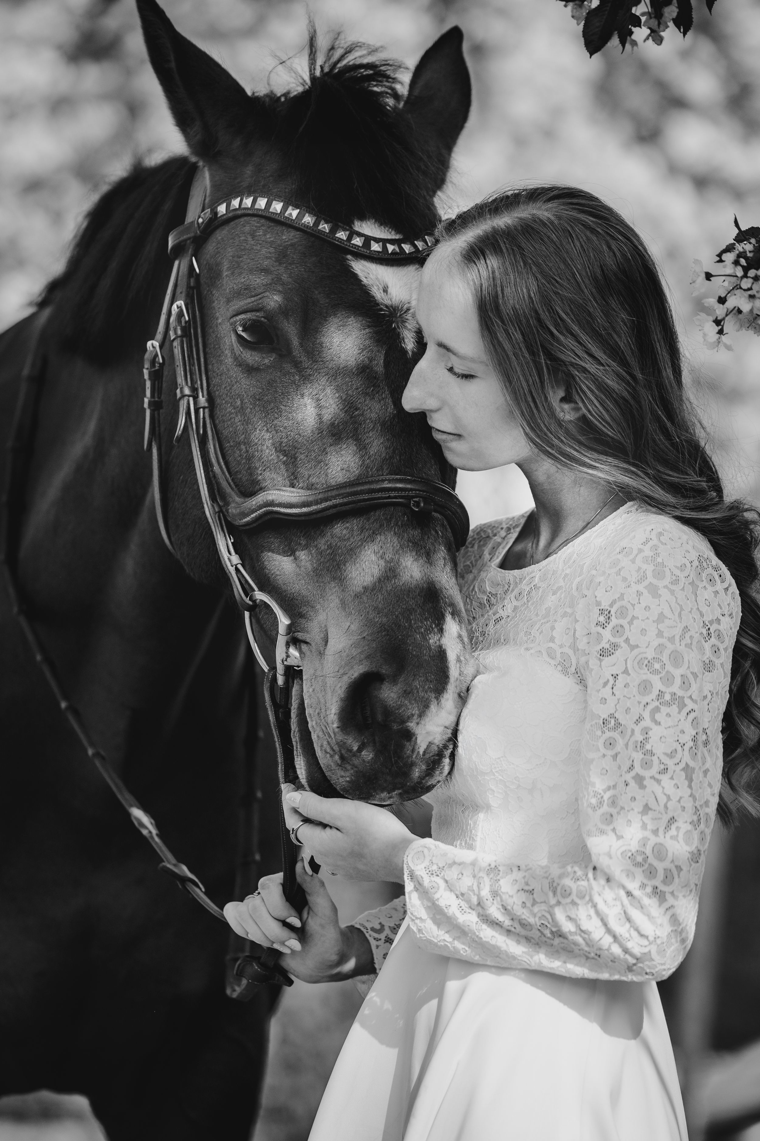 Young woman in white lace dress tenderly embracing a dark bay horse wearing a bridle, black and white.