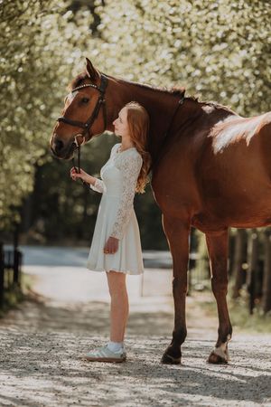A young redhead woman in a white lace dress stands beside a chestnut horse on a sunlit tree-lined path.