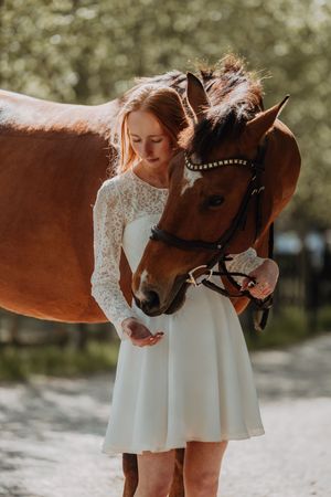 A young woman in a white lace dress gently feeds a chestnut horse outdoors in dappled sunlight.