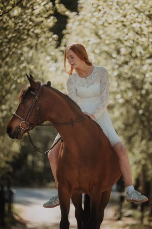 Redhead woman in white lace dress riding a chestnut horse through a blooming tree-lined path.