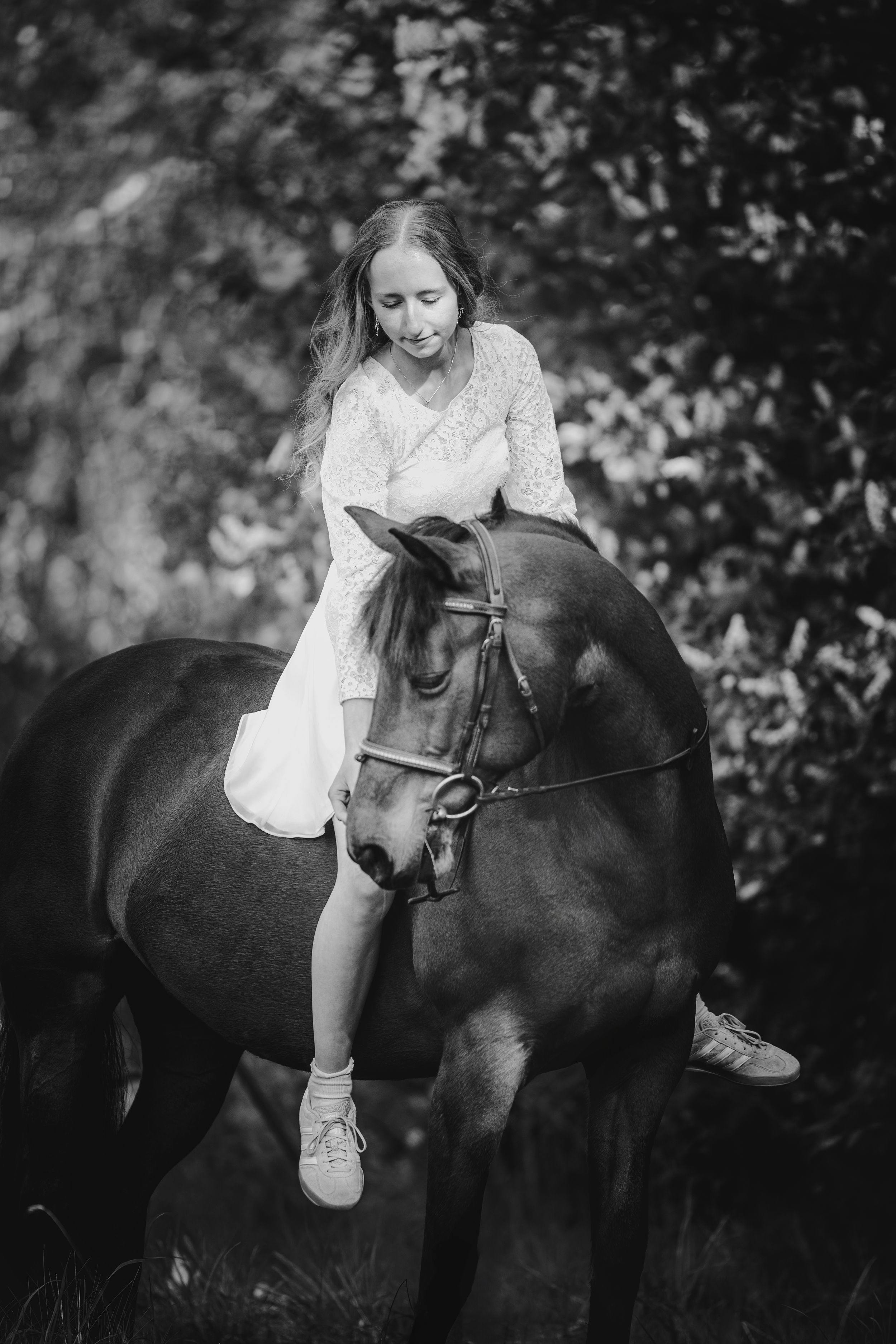Young woman in white dress sitting bareback on a dark horse in a lush green setting, black and white.