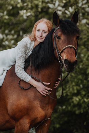 Red-haired woman in white lace dress leaning affectionately on a bay horse in a green outdoor setting.