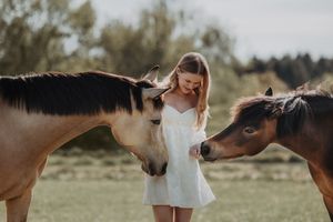 Young woman in white dress standing between two horses in a green field, gently petting them on a sunny day.