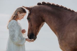 Young woman in white lace dress gently holding a chestnut horse's face against a soft sky background.