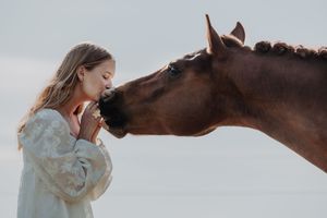 Young blonde girl shares a tender moment nose-to-nose with a chestnut horse against a cloudy sky.