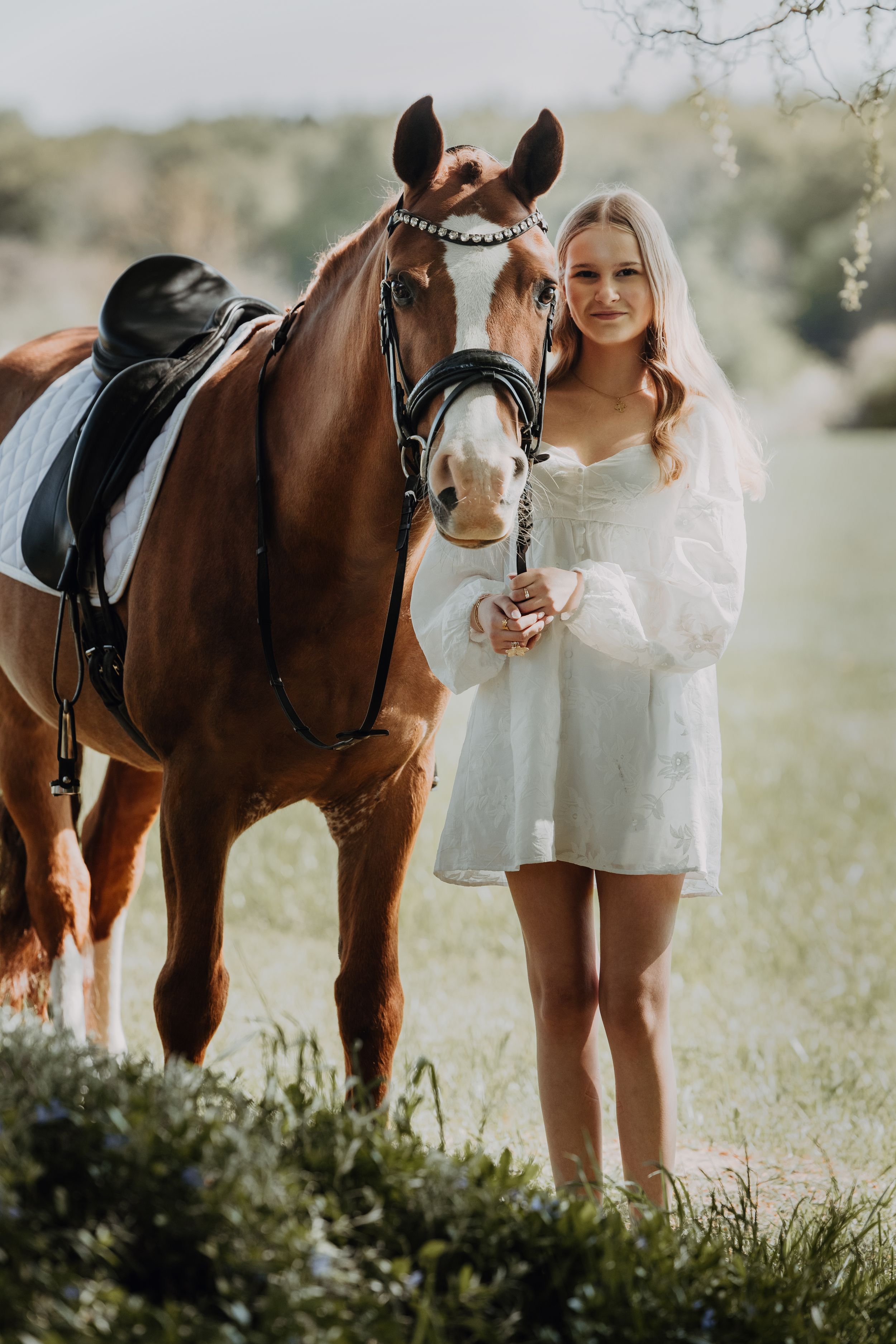 Blonde woman in white dress standing beside saddled chestnut horse in green outdoor setting.