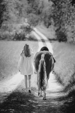 Young girl in white dress walking beside a horse down a sunlit country path, black and white photo.