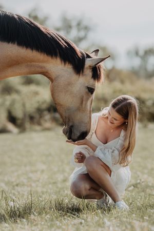 Young woman in white dress crouching to feed a buckskin horse in an open grassy field.