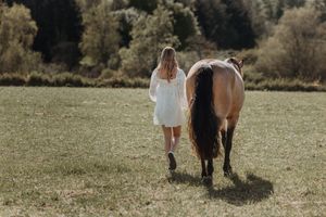 Woman in white dress walking alongside a buckskin horse in a green meadow with trees in background.