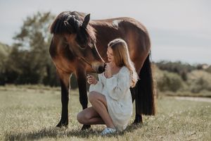 Young woman in white dress kneeling beside a brown horse in a sunny green field.