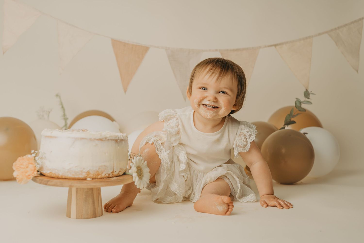 Baby in white outfit crawls on beige backdrop with neutral toned decorations and bunting in studio setting.