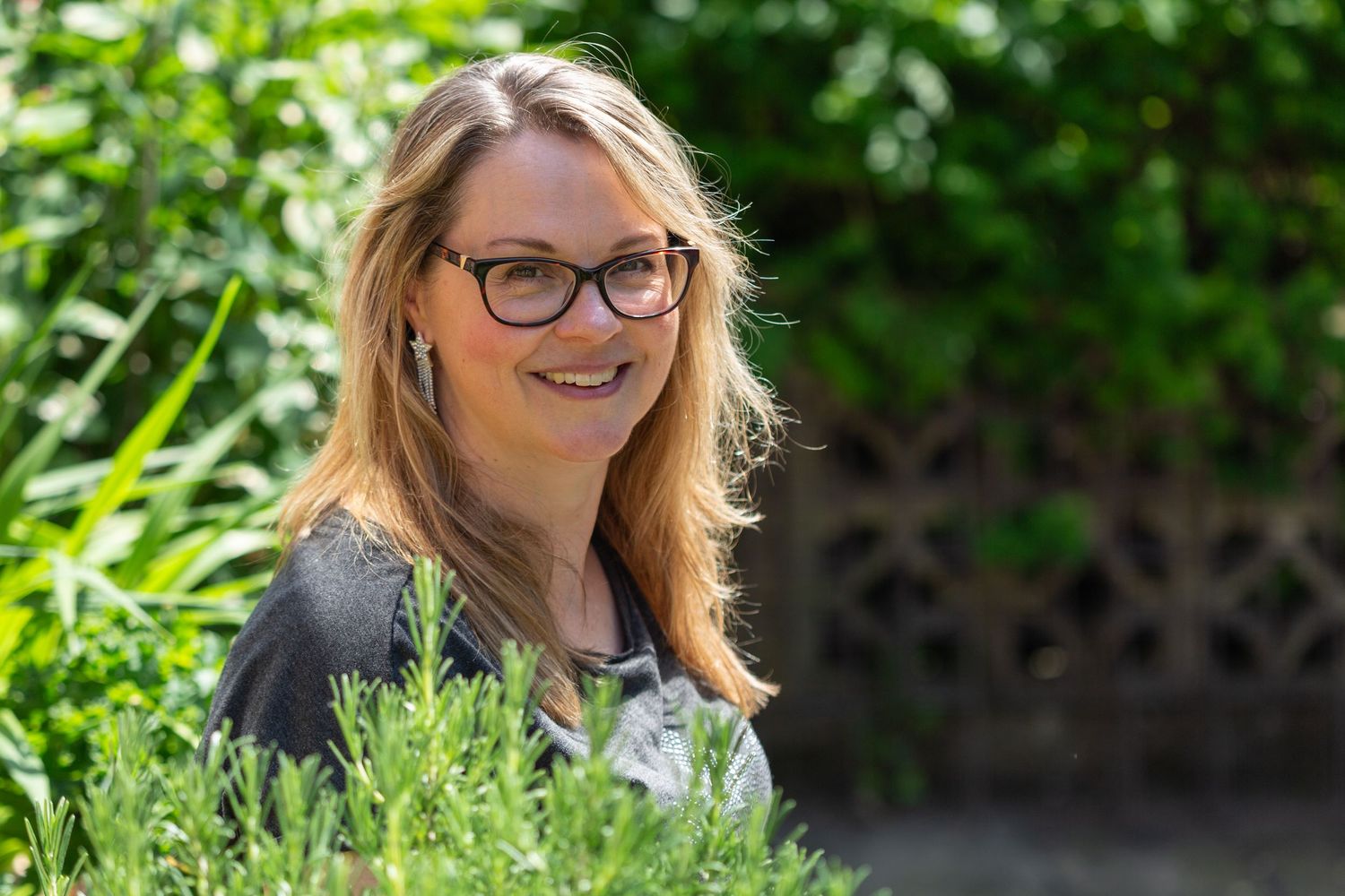 A person with blonde hair and glasses smiles outdoors in a sunny garden setting with lush green foliage.