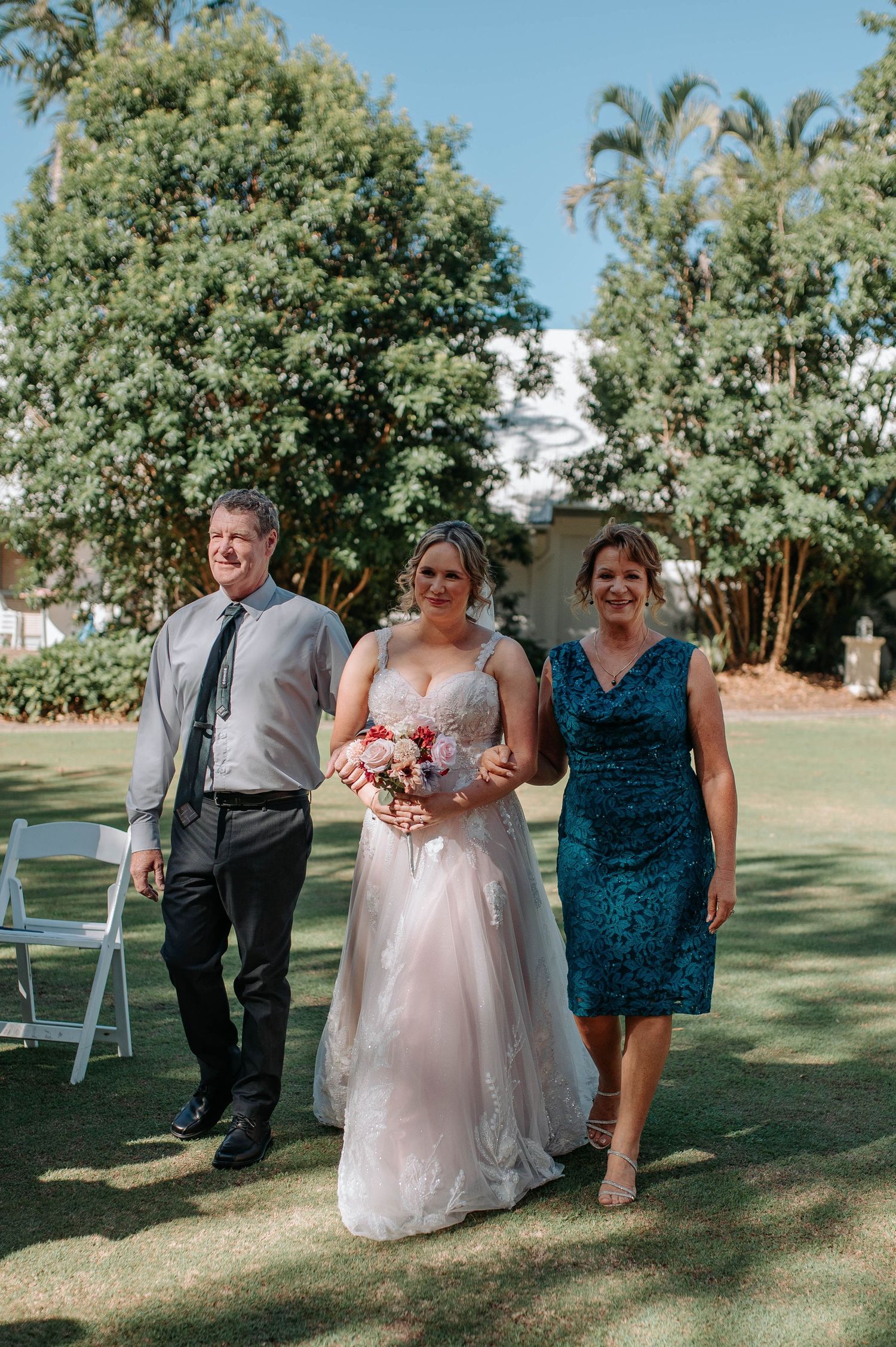 A wedding processional walks down a grassy outdoor aisle during a sunny summer ceremony.
