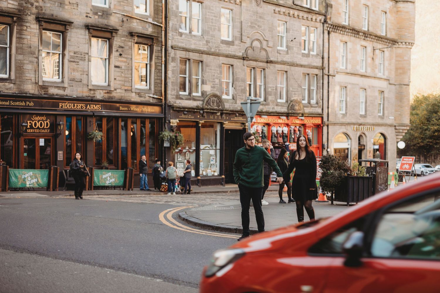 People walking on a charming street corner with historic stone buildings and shops in Edinburgh, Scotland at sunset.