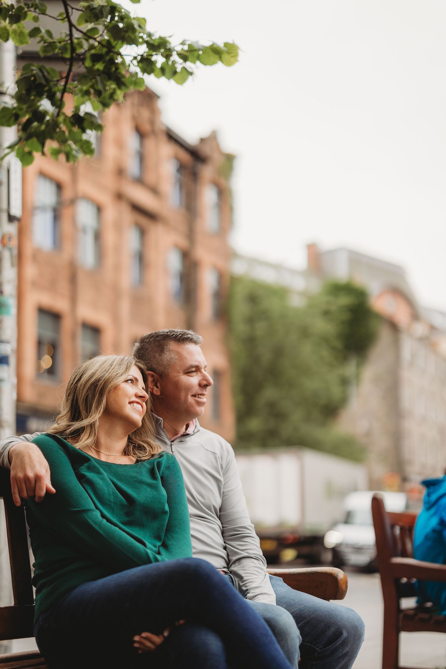Couple relaxing on outdoor bench in urban setting with brick buildings and greenery in background.