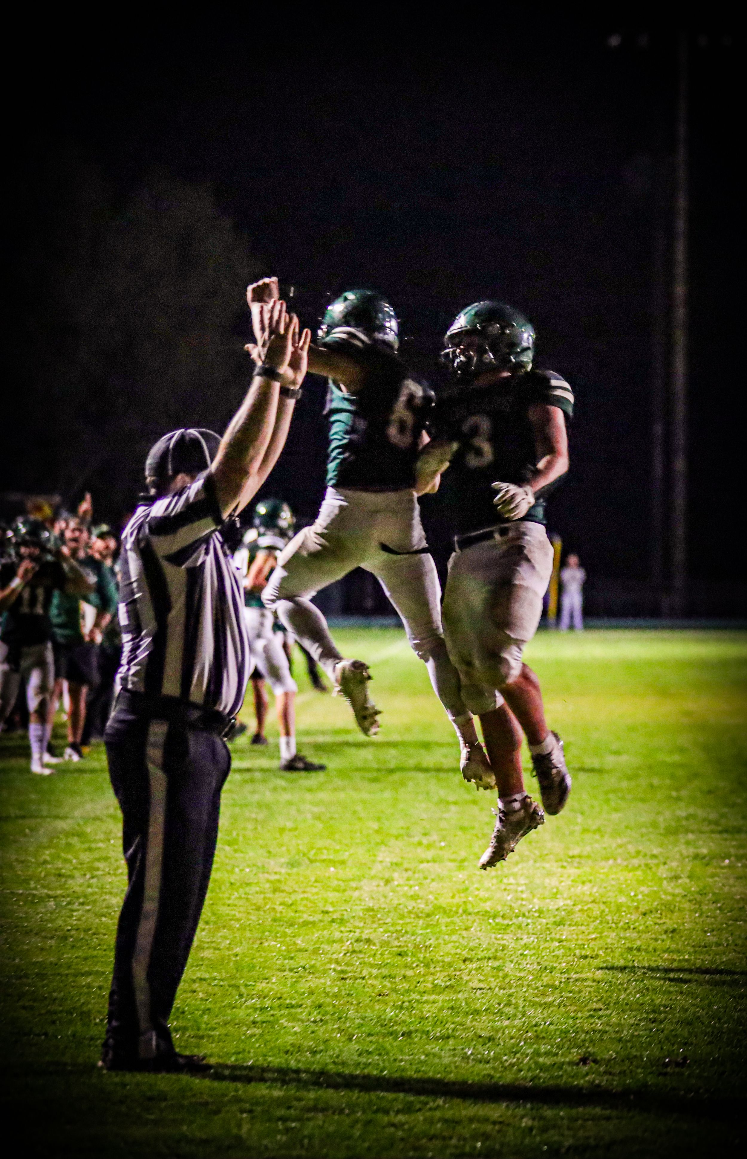 Two youth football players in green uniforms celebrate with a jump near a referee on night field.