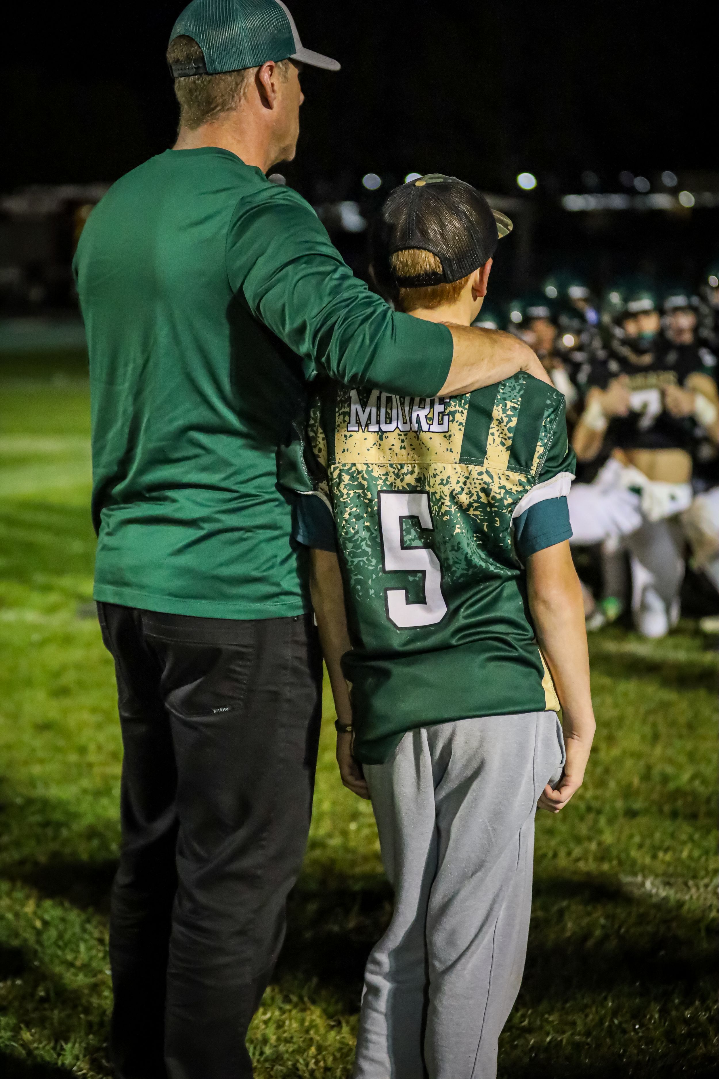 Coach places arm around youth player wearing Moore number 5 green jersey on football sideline at night.