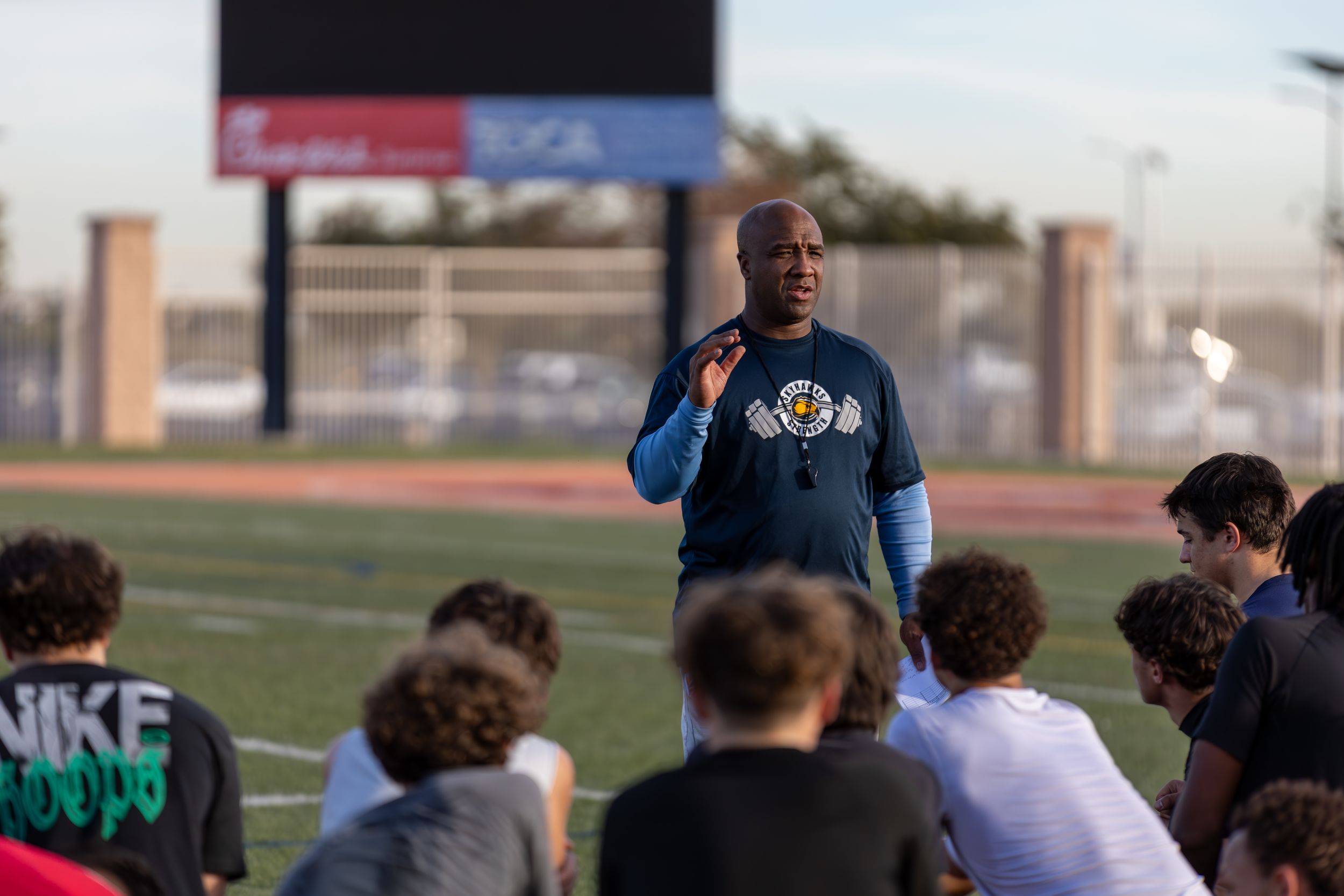 A coach addresses a group of young football players seated on a field during an Exact Sports combine.