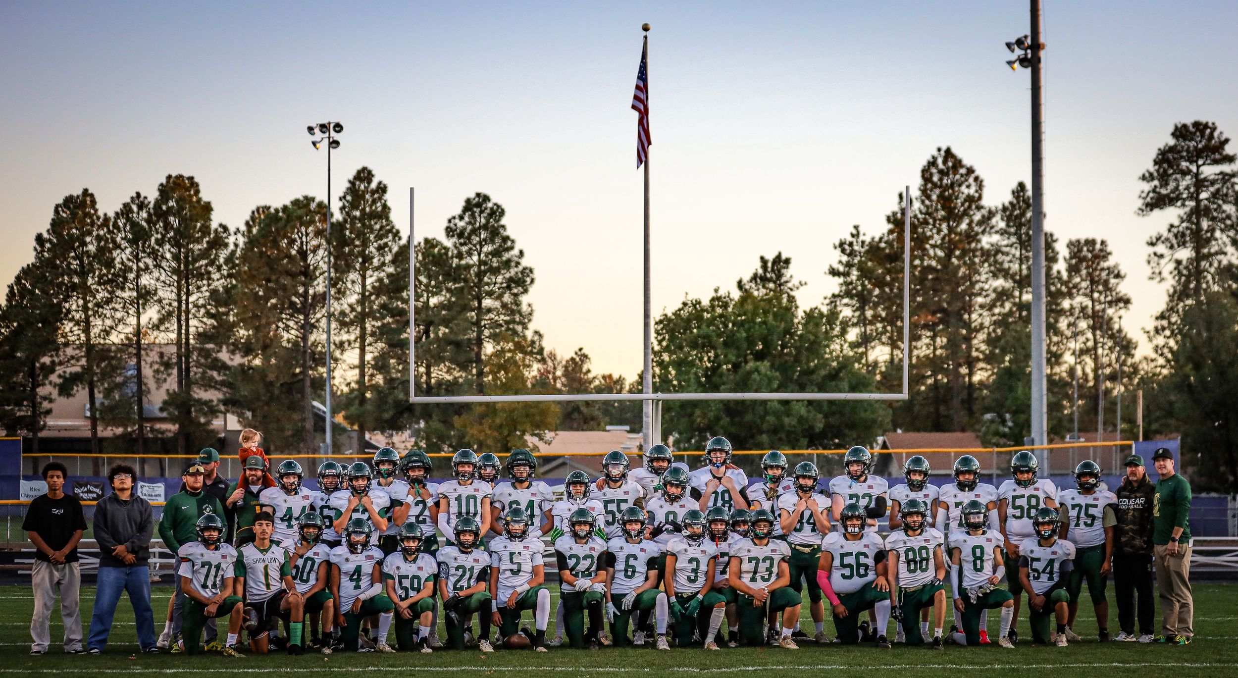High school football team poses for group photo in front of American flag at outdoor stadium during dusk.