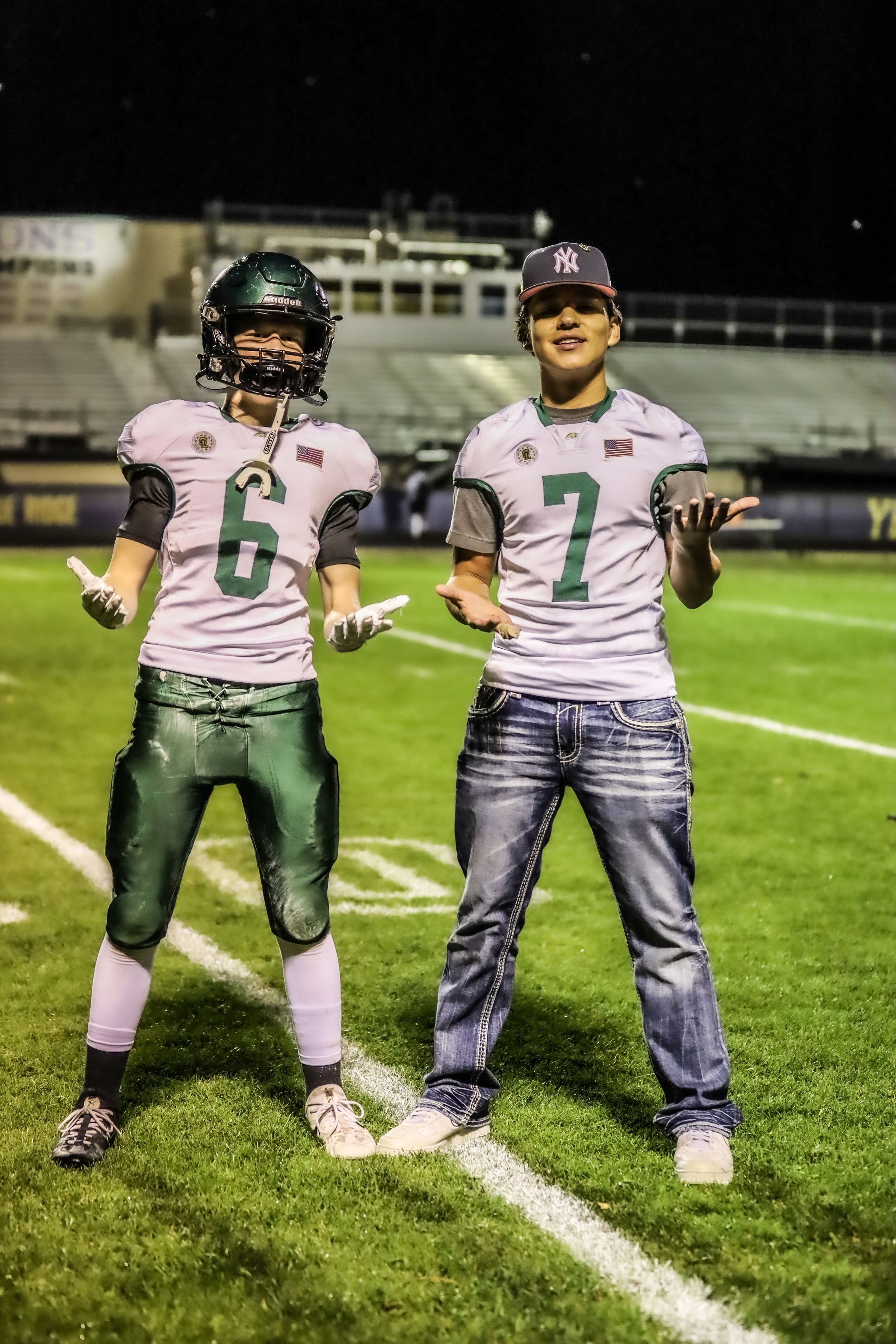Two youth football players wearing numbers 6 and 7 in green and white uniforms pose on field at night.