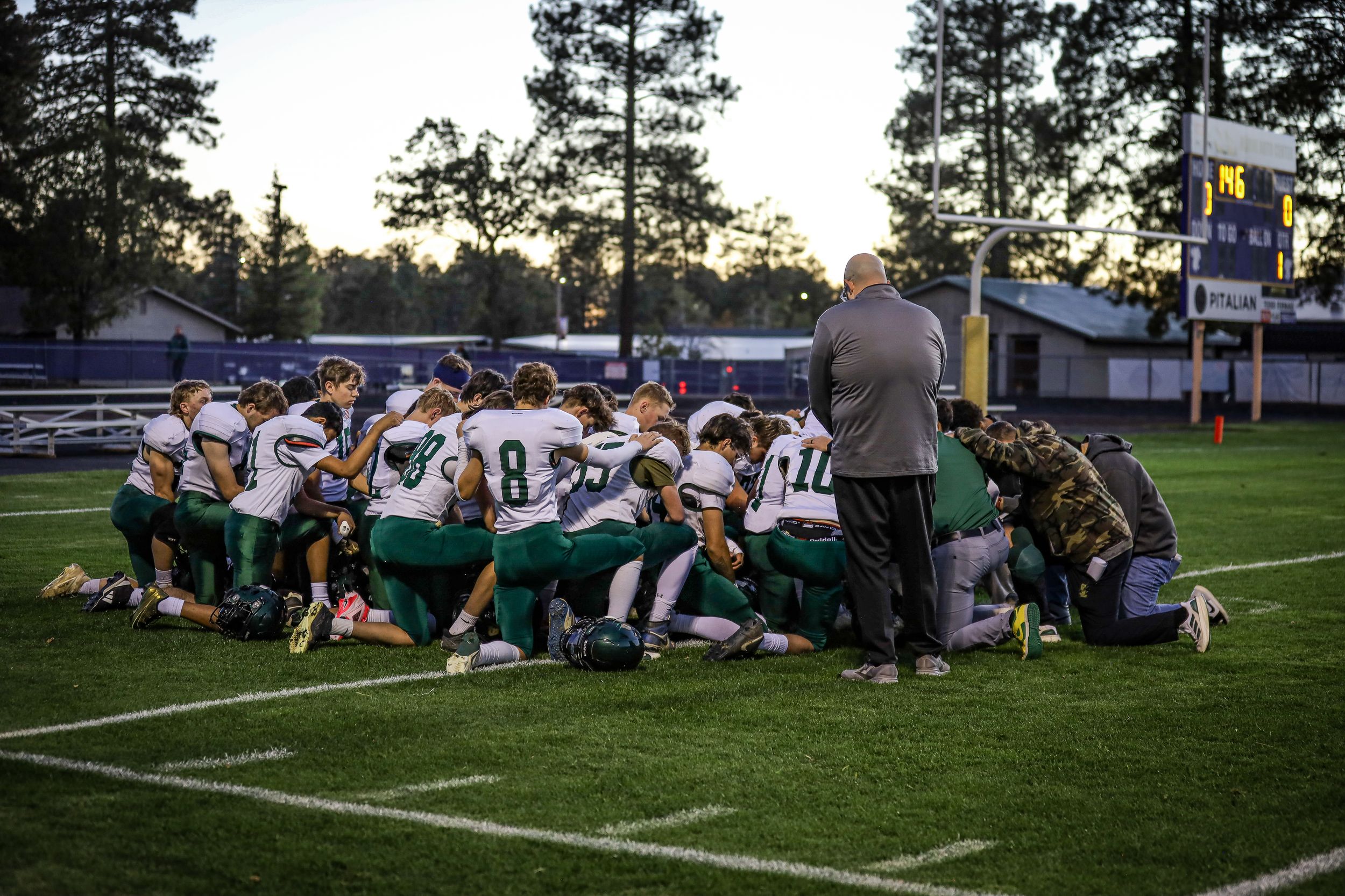 Youth football players huddle in prayer with coach on field before game at dusk.
