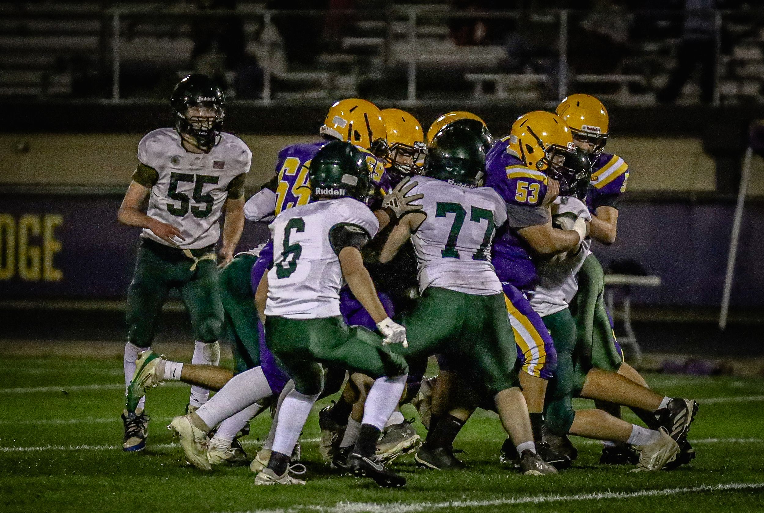 Youth football players in green and gold uniforms clash during an intense tackle play on field.